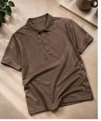 A brown short-sleeve polo shirt with a three-button placket, laid flat on a textured beige surface with a plant leaf and a ceramic mug in the background.