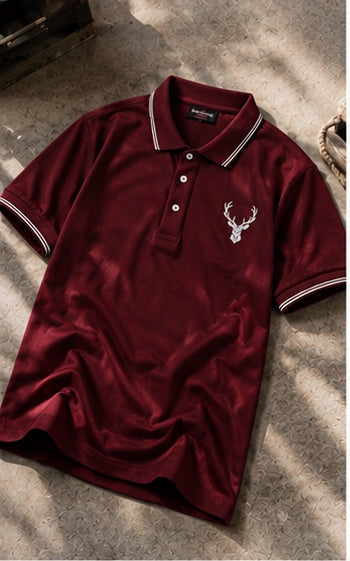 A short-sleeve polo shirt in deep burgundy, laid flat on a textured stone surface with dappled sunlight and shadows. The shirt features a white embroidered deer head logo on the left chest and white contrast piping along the collar and sleeve cuffs.