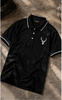 A black short-sleeve polo shirt with white piping on the collar and sleeve cuffs, featuring a white embroidered deer head logo on the left chest. The shirt is laid flat on a textured gray stone surface with soft shadows.