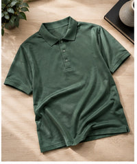 A dark green short-sleeve polo shirt laid flat on a light wood surface, with a small plant and a book visible in the corners as styled props.