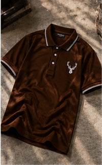 A brown short-sleeve polo shirt with white contrast piping on the collar and sleeve cuffs, featuring a white embroidered deer head logo on the left chest. The shirt is laid flat on a textured concrete surface with dappled sunlight and shadows.