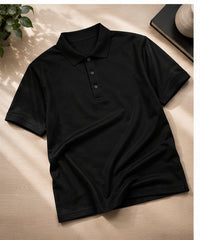 A black short-sleeve polo shirt laid flat on a light beige surface with soft shadows, alongside a book, a small vase, and a leafy plant in the corners.