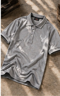 A gray short-sleeve polo shirt with a small white deer head logo on the left chest, laid flat on a textured concrete surface with dappled sunlight and shadows.