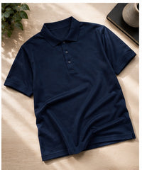 A navy blue short-sleeve polo shirt laid flat on a light beige surface with soft shadows, surrounded by small decorative props including a plant leaf and a ceramic cup.