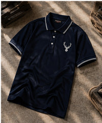 A navy blue short-sleeve polo shirt with white striped collar and cuffs, featuring a white embroidered deer head logo on the left chest, laid flat on a textured concrete surface with dappled sunlight.