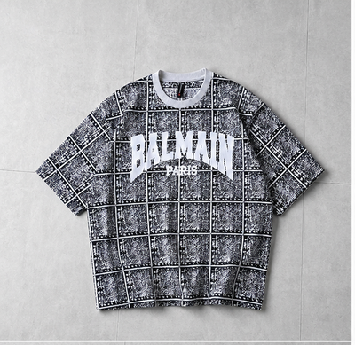 A Balmain short-sleeve t-shirt with a black and white grid pattern and a white 'BALMAIN PARIS' logo on the chest, laid flat on a gray tiled background.