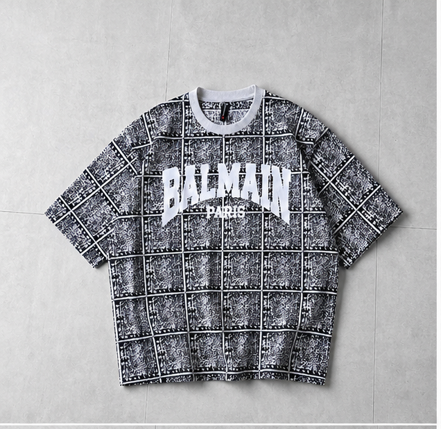 A Balmain short-sleeve t-shirt with a black and white grid pattern and a white 'BALMAIN PARIS' logo on the chest, laid flat on a gray tiled background.
