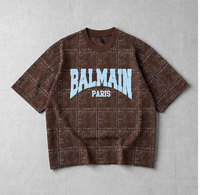 A brown Balmain Paris short-sleeve t-shirt with a textured grid pattern and light blue 'BALMAIN PARIS' text on the front, laid flat on a gray background.