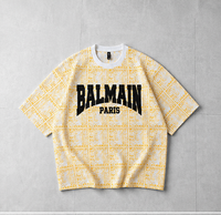 A Balmain Paris short-sleeve t-shirt with a yellow and white monogram pattern, featuring a large black 'BALMAIN PARIS' logo on the chest, laid flat on a gray tiled background.