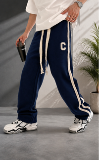 A person wearing navy blue sweatpants with cream drawstrings and a cream letter 'C' on the thigh, styled with a white t-shirt and chunky white sneakers in a modern indoor setting.