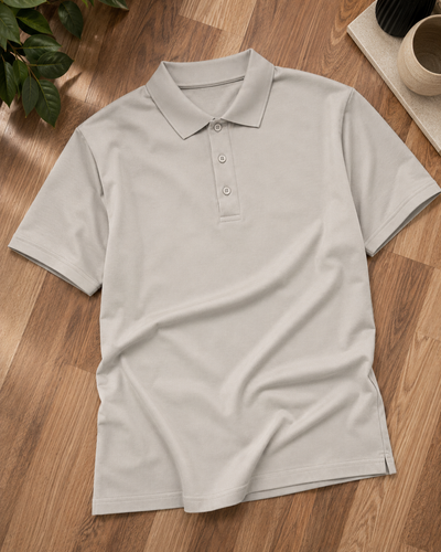 A light beige short-sleeve polo shirt laid flat on a wooden floor, with a green plant and a ceramic cup visible in the corners as styling props.