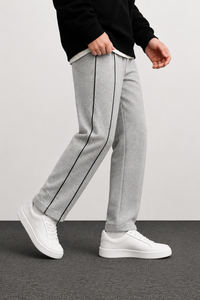 A person wearing light gray heather sweatpants with a black side stripe, paired with a black top and white sneakers, shown from the waist down against a light gray wall and dark gray floor.