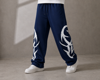 A person wearing navy blue sweatpants with large white graphic prints on both legs, paired with white sneakers, standing against a textured gray studio wall.