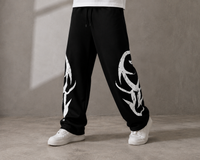 Black sweatpants with large white graphic prints on both legs, worn by a model with white sneakers against a textured gray studio background.
