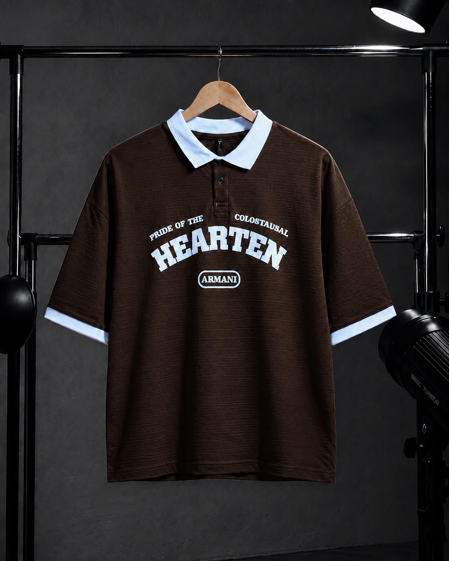 A dark brown short-sleeve polo shirt with a light blue collar and sleeve cuffs, featuring a white graphic print on the front that reads 'PRIDE OF THE COLOEBSTRAIC HEARTEN' and 'ARMANI'. The shirt is displayed on a wooden hanger against a dark gray background with studio lighting equipment visible on the sides.