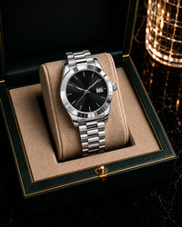 A luxury stainless steel wristwatch with a black dial and date window, presented on a beige cushion inside an open dark green and gold watch box.