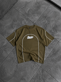 An olive green short-sleeve t-shirt with white contrast piping and a white 'SNTK' logo on the chest, laid flat on a dark gray tiled floor.