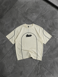 A cream-colored short-sleeve t-shirt with black piping details and a black 'Skater' graphic on the chest, laid flat on a dark gray tiled floor.