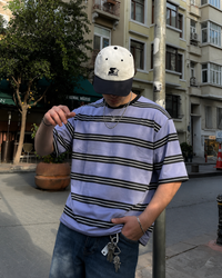 A man wearing a light purple and black horizontally striped t-shirt, blue jeans, and a white baseball cap with a black brim, standing on a city street.