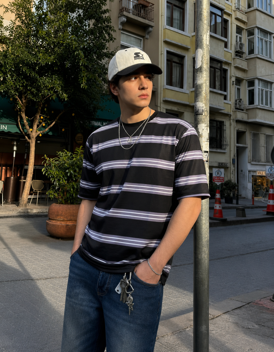 A man wearing a black short-sleeve t-shirt with thin white and purple horizontal stripes, blue jeans, a white and black baseball cap, and silver necklaces, standing on a city street.