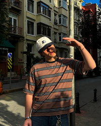 A man wearing a brown, black, and white striped t-shirt, blue jeans, a white baseball cap, and sunglasses stands on a city street in front of apartment buildings.