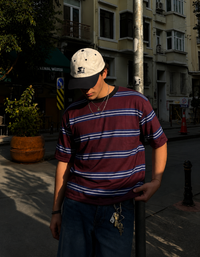 A man wearing a burgundy t-shirt with thin blue and white horizontal stripes, a cream baseball cap, and dark blue jeans, standing on a city street in warm sunlight.
