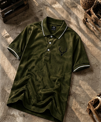 An olive green short-sleeve polo shirt with white piping on the collar and cuffs, featuring a black embroidered deer head logo on the left chest. The shirt is laid flat on a textured brown surface with rustic wooden props in the background.