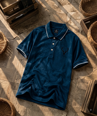 A dark blue short-sleeve polo shirt with white piping on the collar and cuffs, featuring a black embroidered deer logo on the left chest. The shirt is laid flat on a rustic wooden floor surrounded by woven baskets.