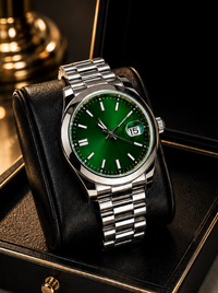 A silver stainless steel wristwatch with a vibrant green dial and a date window displaying '15', resting on a black leather watch cushion inside a black presentation box.
