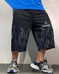 Black mid-thigh shorts with a gray flame print and a white Jordan logo on the left thigh, worn by a model with gray and blue sneakers.