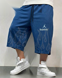 A person wearing blue cropped sweat shorts with white flame and Jumpman graphics, paired with white high-top sneakers, standing against a plain light gray wall.