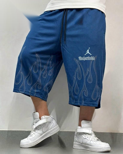 A person wearing blue cropped sweat shorts with white flame and Jumpman graphics, paired with white high-top sneakers, standing against a plain light gray wall.