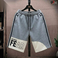 Gray heathered athletic shorts with black side stripes, a white contrast panel on the lower left leg reading 'BESTRONG FEAR', and a black drawstring, hanging on a black hanger against a brown textured backdrop.