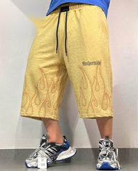 A person wearing loose yellow shorts with black drawstrings, orange flame outlines, and the word 'Uncomfortable' printed near the right thigh, paired with white and blue sneakers against a plain indoor wall.