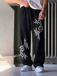 A person wearing black drawstring sweatpants with large white tribal-style graphic prints on both legs, paired with a white t-shirt and white sneakers, standing in an indoor setting.