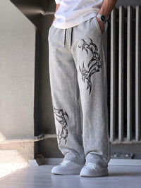 A person wearing light gray heather sweatpants with black tribal-style graphic prints on the legs, paired with a white t-shirt and white sneakers, standing in an indoor setting.