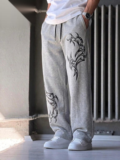 A person wearing light gray heather sweatpants with black tribal-style graphic prints on the legs, paired with a white t-shirt and white sneakers, standing in an indoor setting.