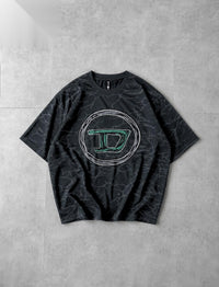 A black short-sleeve t-shirt with a subtle gray marble pattern, featuring a large circular graphic on the chest with a stylized letter 'D' outlined in green and embellished with rhinestones, laid flat on a gray tiled surface.