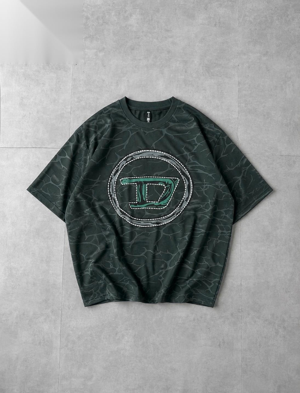 A dark green short-sleeve t-shirt with a subtle marble pattern, featuring a large circular graphic on the chest with a stylized letter 'D' outlined in green and bordered with rhinestones, laid flat on a gray tiled background.