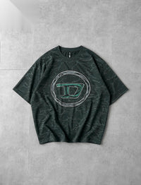 A dark green short-sleeve t-shirt with a subtle marble pattern, featuring a large circular graphic on the chest with a stylized letter 'D' outlined in green and bordered with rhinestones, laid flat on a gray tiled background.