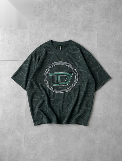 A dark green short-sleeve t-shirt with a subtle marble pattern, featuring a large circular graphic on the chest with a stylized letter 'D' outlined in green and bordered with rhinestones, laid flat on a gray tiled background.
