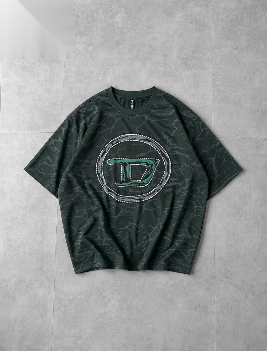 A dark green short-sleeve t-shirt with a subtle marble pattern, featuring a large circular graphic on the chest with a stylized letter 'D' outlined in green and bordered with rhinestones, laid flat on a gray tiled background.