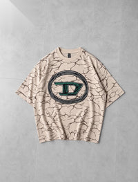 A beige short-sleeve t-shirt with a dark brown cracked earth pattern and a large circular graphic on the chest featuring a stylized letter 'D' in green and black, laid flat on a gray concrete background.