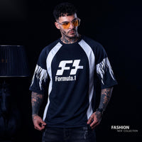 A man wearing a black and white Formula.1 raglan t-shirt with flame graphics on the sleeves, styled with orange-tinted sunglasses against a dark studio background.