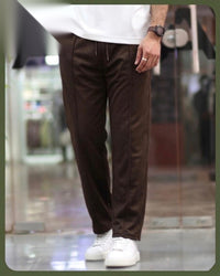 A person wearing dark brown drawstring trousers, a white t-shirt, and white sneakers, standing in a blurred indoor environment.