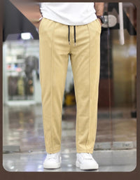 A person wearing beige drawstring trousers with a white t-shirt and white sneakers, shown from the waist down in a blurred indoor setting.