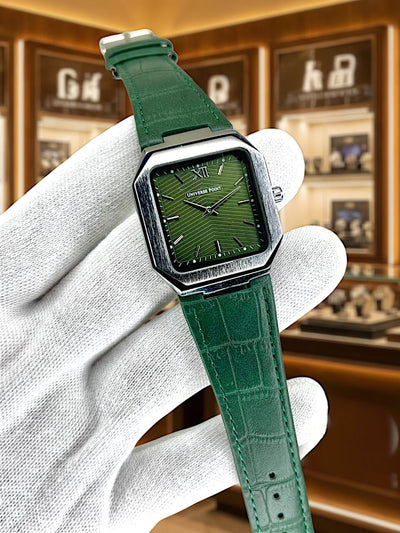 Green wristwatch with a detailed face held by a gloved hand against a blurred background.