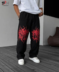 A person wearing black sweatpants with large red graphic prints on the thighs, paired with a white t-shirt and white sneakers, standing on a dark wood floor.