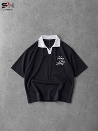 A black short-sleeve polo shirt with a white collar and white script embroidery on the left chest, laid flat on a gray tiled surface.