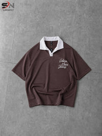 A short-sleeve polo shirt in dark brown with a white collar and white script embroidery on the chest, laid flat on a gray tiled surface.
